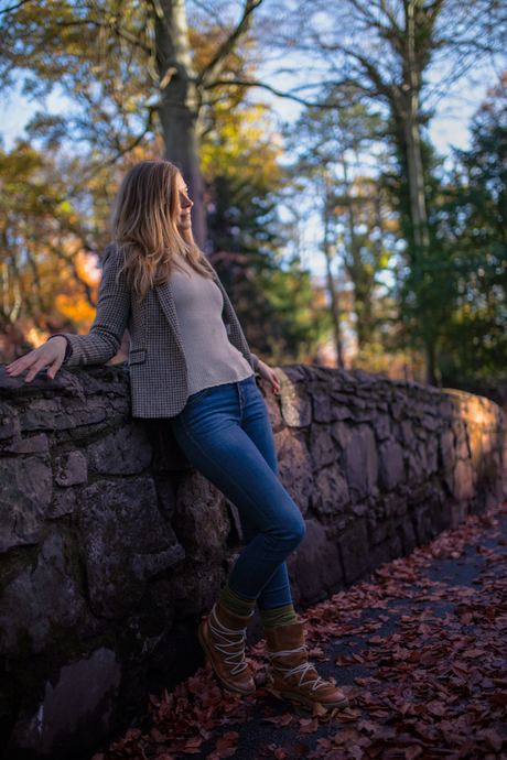 Fashion photo of a woman in a grey top and jeans leaning on a stone wall in autumn
