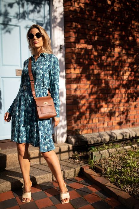 Fashion photo of a woman in a blue floral shirt dress with a crossbody bag beside a red brick wall