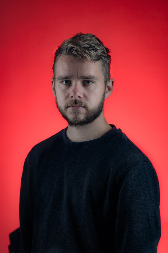 Headshot of a man in a navy jumper against a red background