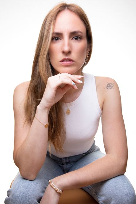Portrait of a woman seated in a white vest top and jeans, hand at her chin